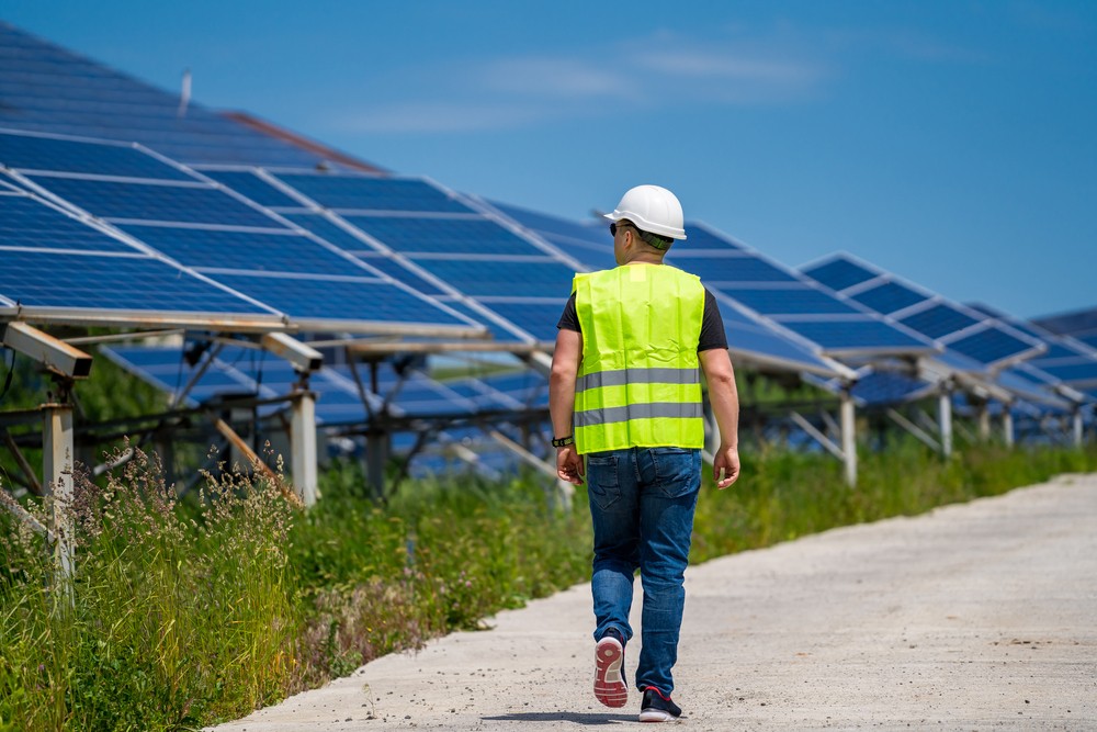 An engineer wearing a hard hat and vest walking in front of solar panels 