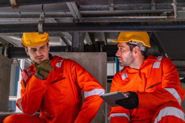 Two Engineers In Orange Coveralls Examine Industrial Pipes